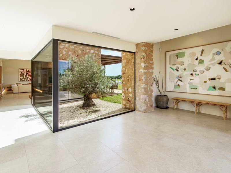 Pasillo moderno con grandes ventanales que muestran un patio interior con un olivo y paredes de piedra natural.
