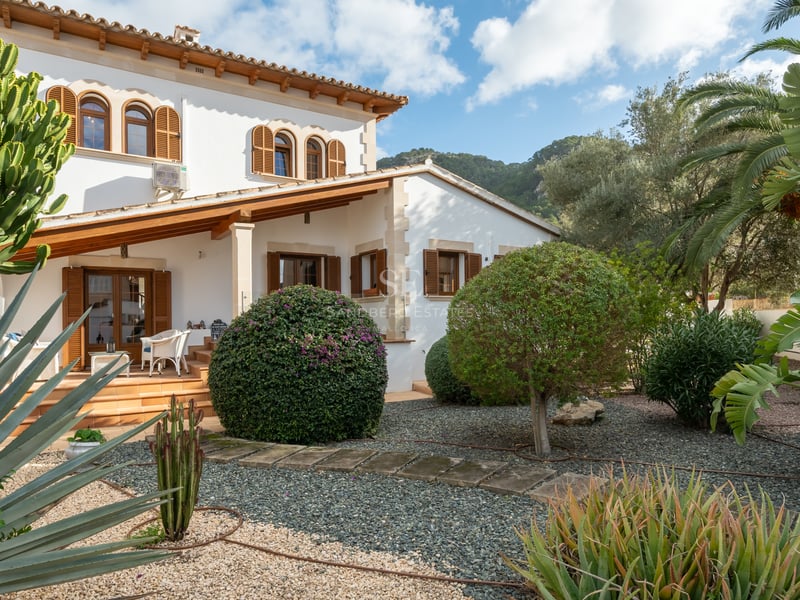 Villa majorquine blanche avec volets en bois et jardin de cactus sur fond de montagnes.