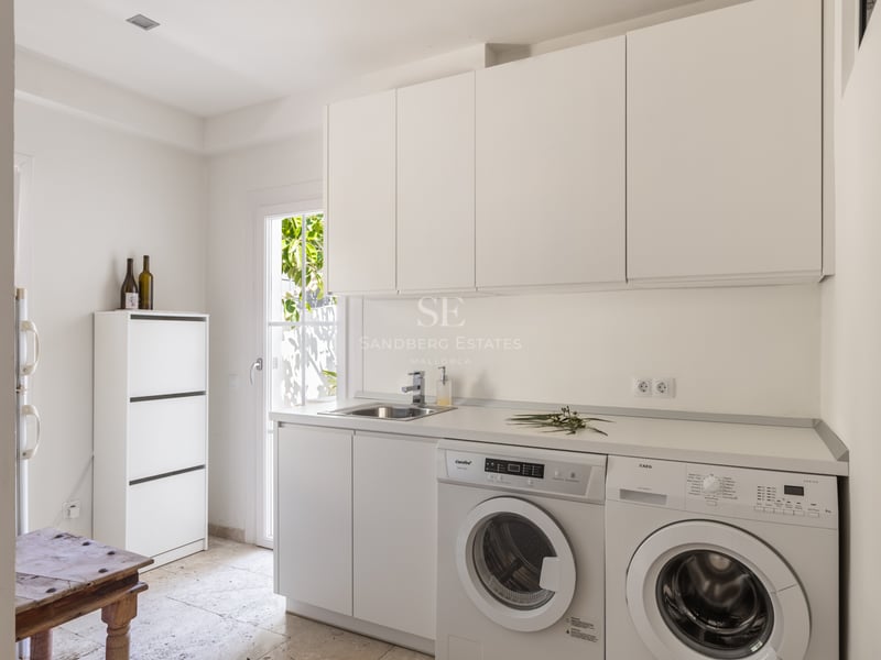 Buanderie blanche avec lave-linge, sèche-linge et évier en inox sur un sol en pierre naturelle claire.