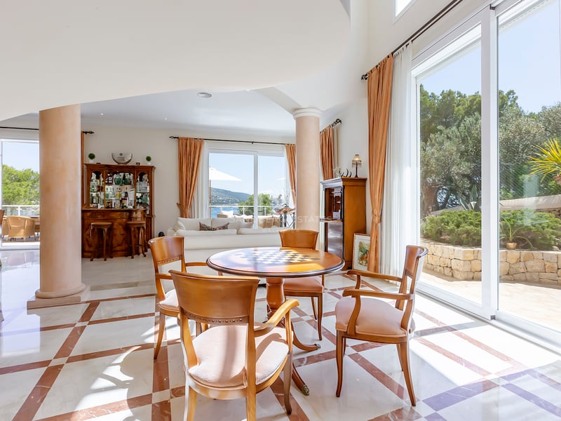 Spacieux salon avec sol en marbre, table en bois, colonnes et grandes fenêtres donnant sur un jardin et la mer.