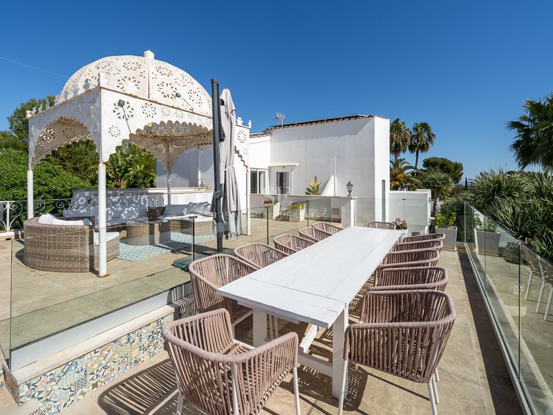Grande terrasse avec table à manger blanche, chaises en osier, barbecue et gazebo orné sous un ciel bleu clair.