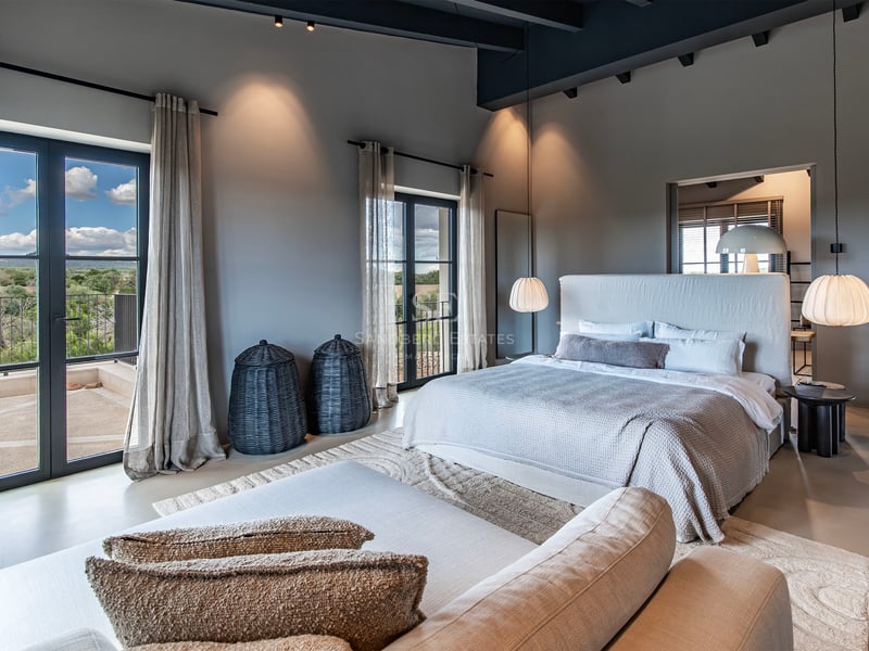 Chambre principale contemporaine avec poutres apparentes, grand lit et portes-fenêtres ouvrant sur un balcon avec vue.