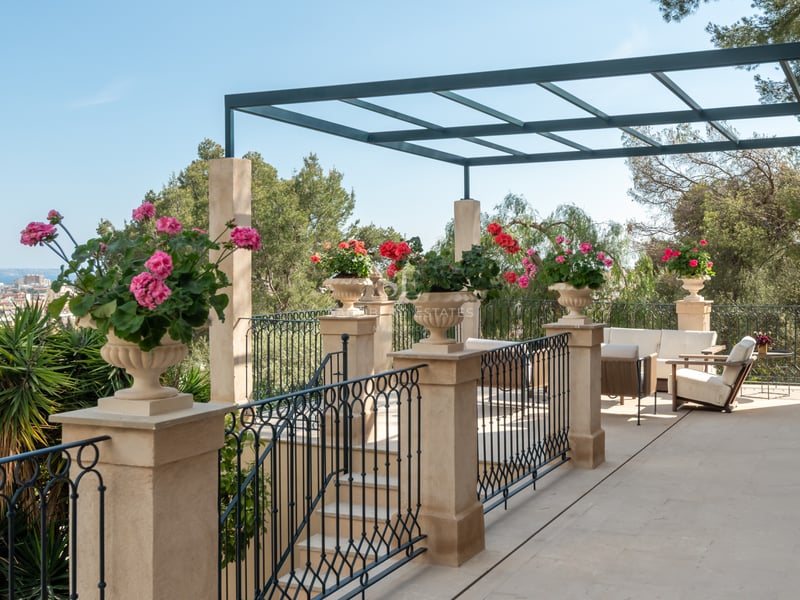 Terrasse en pierre avec garde-corps en fer, pergola métallique et fleurs surplombant la ville et la mer.