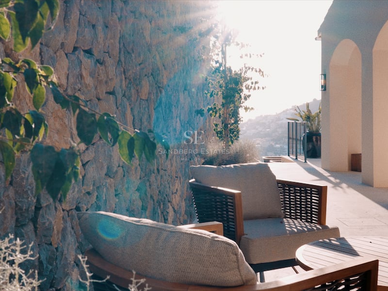 Terrasse ensoleillée avec fauteuils gris élégants, table en bois, mur en pierre brute et piliers blancs arqués.
