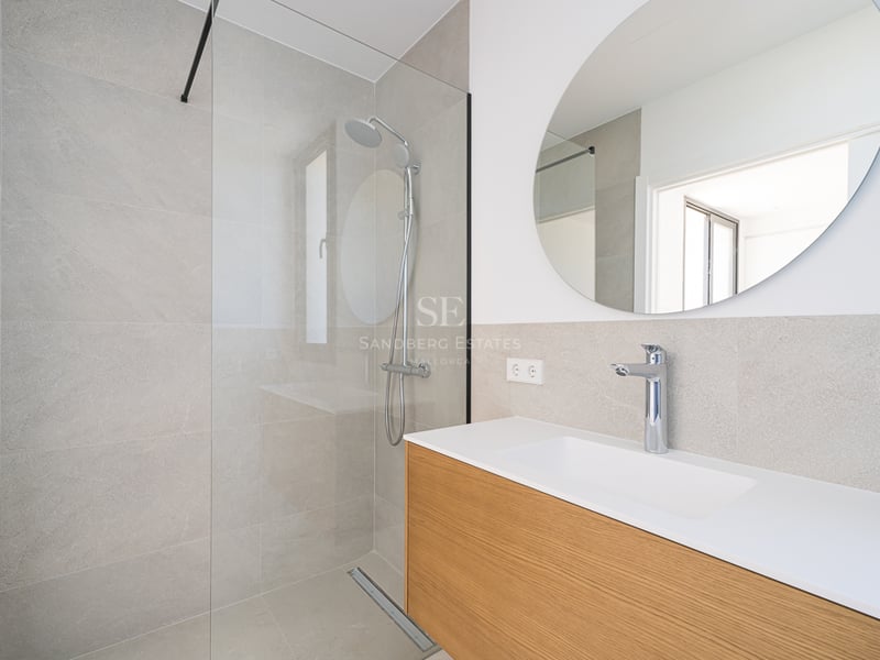 Salle de bain moderne avec douche vitrée, carrelage gris, miroir circulaire et meuble vasque en bois.