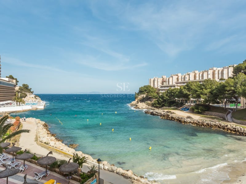 Vue en plongée d'une crique aux eaux turquoise avec une plage, des sentiers en pierre et des immeubles côtiers.