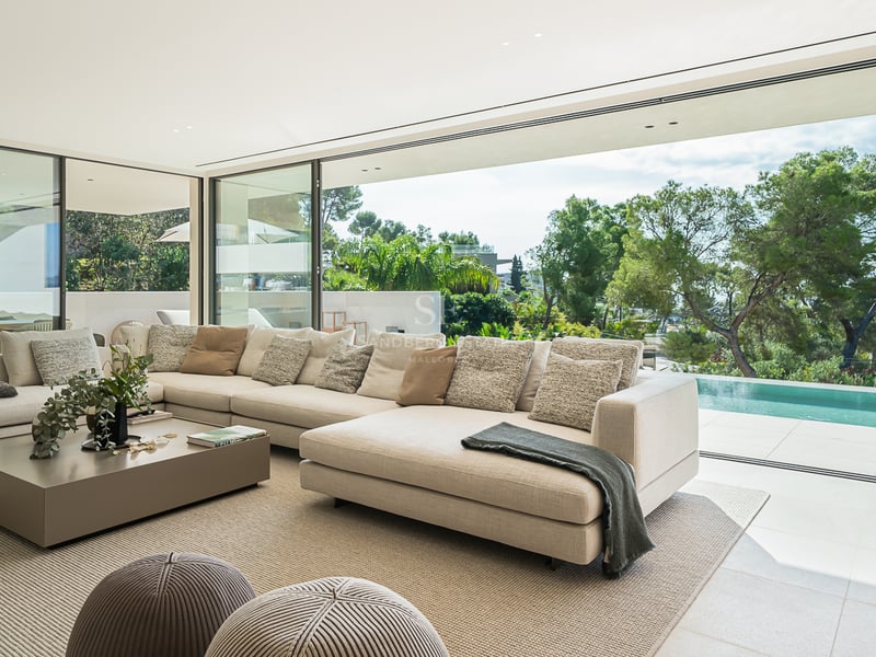 Un salon moderne et lumineux avec un grand canapé d'angle crème donnant sur une piscine privée et des arbres.