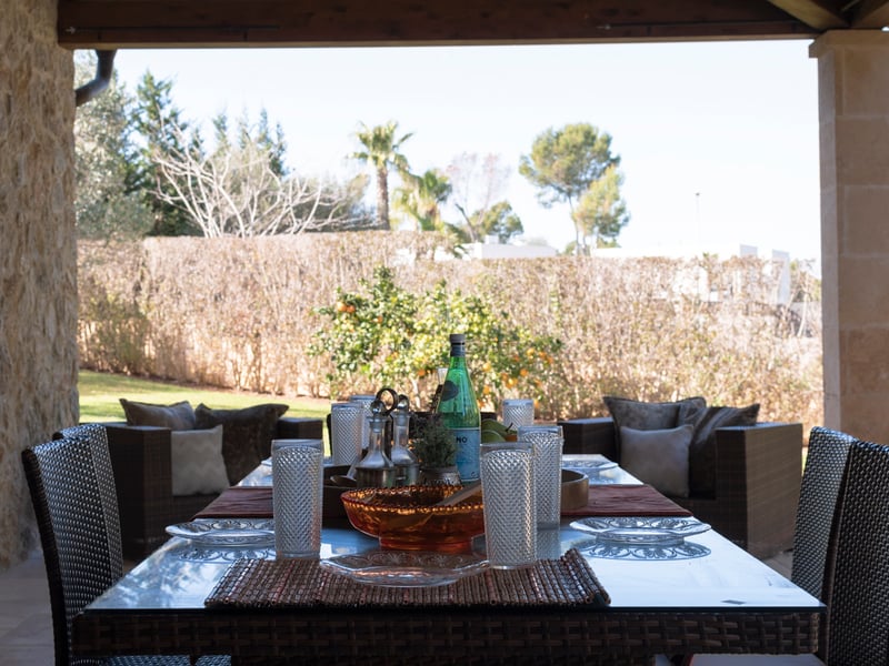 Terrasse méditerranéenne couverte avec salle à manger en osier, murs en pierre naturelle et vue sur un jardin verdoyant.