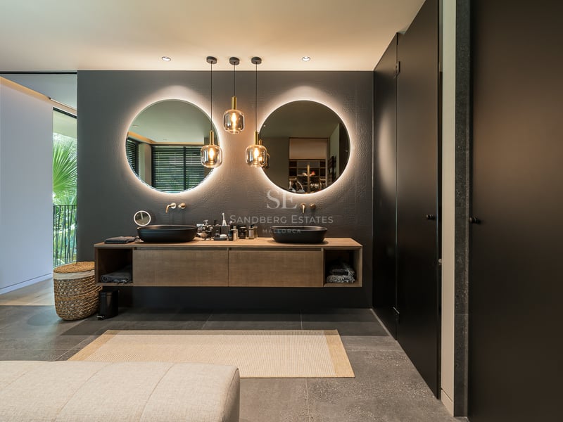 Salle de bain moderne avec deux miroirs ronds éclairés, vasques noires et meuble en bois sur sol en pierre.