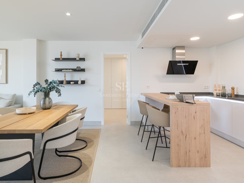 Cocina minimalista blanca con isla de madera y comedor contiguo con sillas de diseño y suelos de piedra clara.