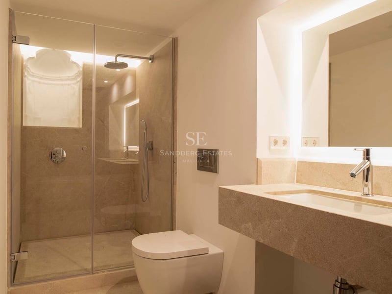 Salle de bain moderne avec douche vitrée, toilettes suspendues et vasque en pierre avec miroir rétroéclairé.