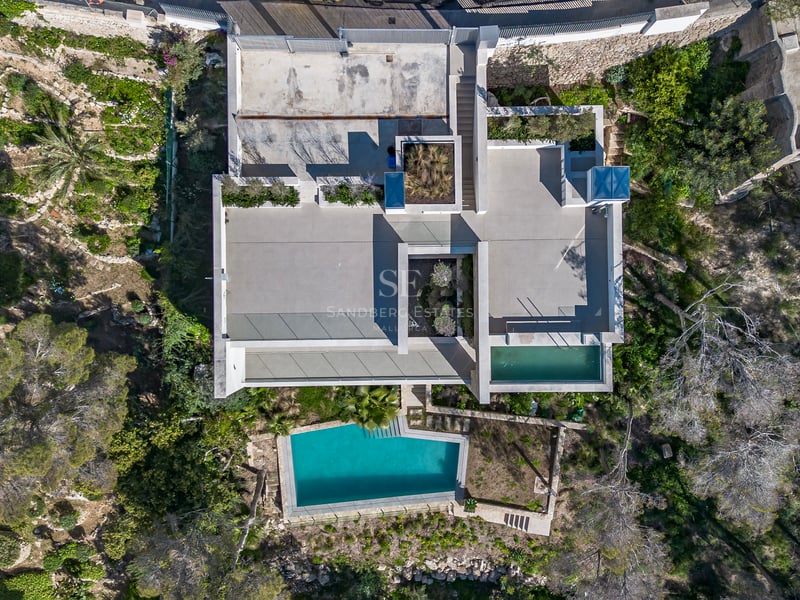 Vue aérienne d'une villa moderne géométrique avec toits plats, terrasses et piscine turquoise cristalline.
