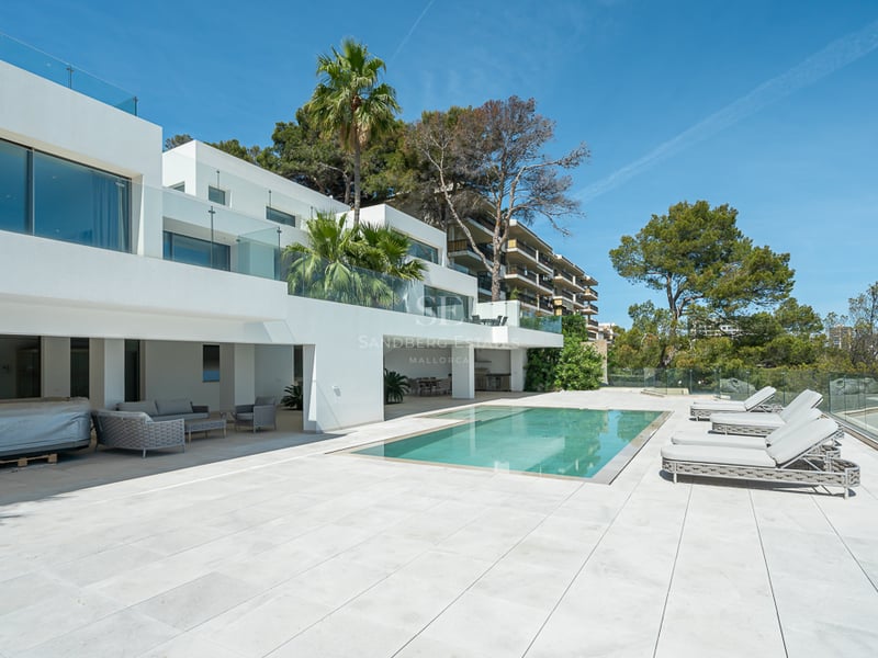 Villa blanche moderne avec piscine rectangulaire, bains de soleil gris et garde-corps en verre sous un ciel bleu.