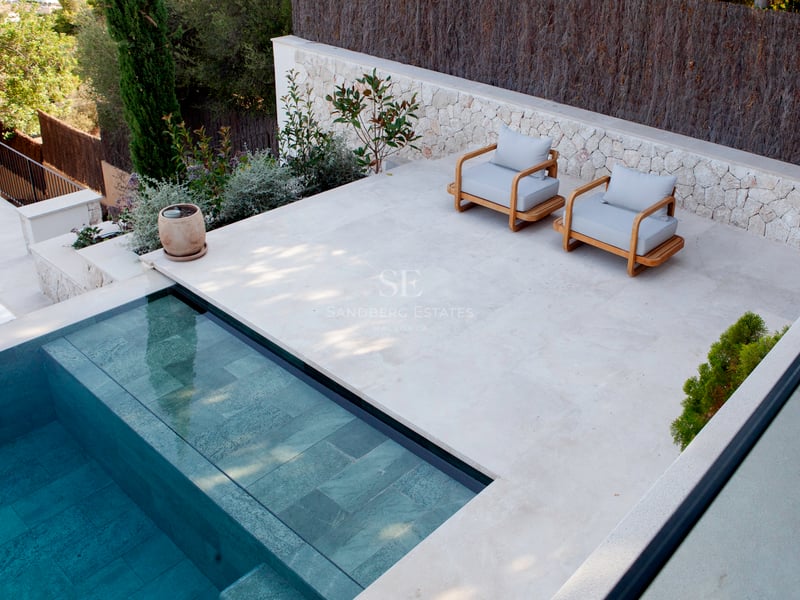 Vue en surplomb d'une piscine carrelée turquoise bordée d'une terrasse en pierre claire avec fauteuils en bois.