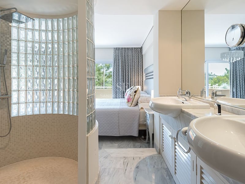 Salle de bains attenante avec double vasque en marbre et douche en briques de verre, ouverte sur la chambre.