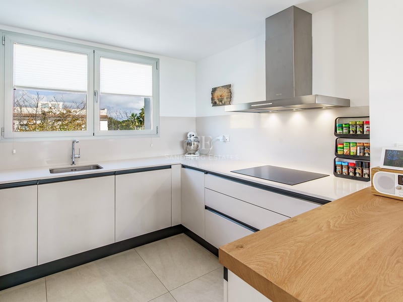 Cuisine blanche moderne en L avec plan de travail en bois, hotte en inox et grande fenêtre.
