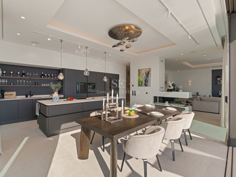 Salle à manger moderne avec table en bois foncé, chaises beiges et cuisine design grise avec grandes baies vitrées.
