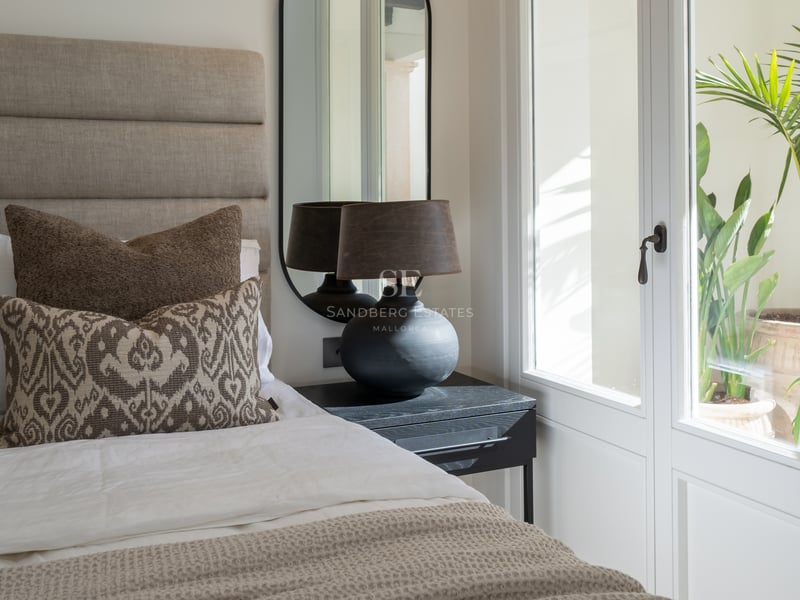 Contemporary bedroom interior with a linen headboard, black designer lamp, and large window overlooking a lush courtyard.