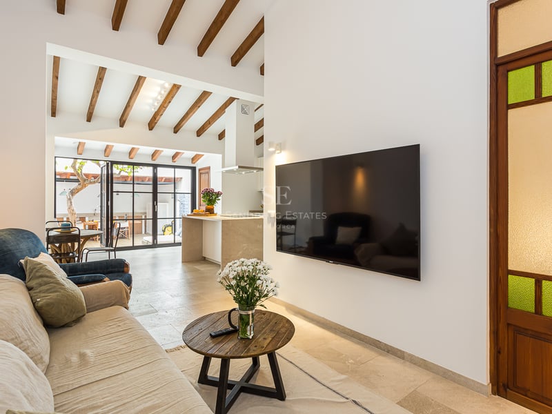 Salon moderne avec canapé beige, poutres en bois au plafond, grand téléviseur et vue sur la cuisine ouverte.