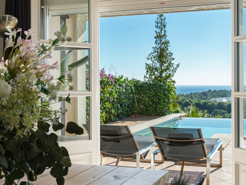 Vue à travers des portes-fenêtres ouvertes sur une terrasse ensoleillée avec chaises longues, piscine et mer.