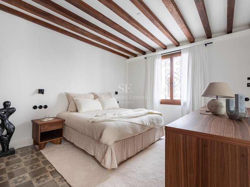 Dormitorio con vigas de madera oscura, suelo de baldosas hidráulicas y una ventana con cortinas blancas.