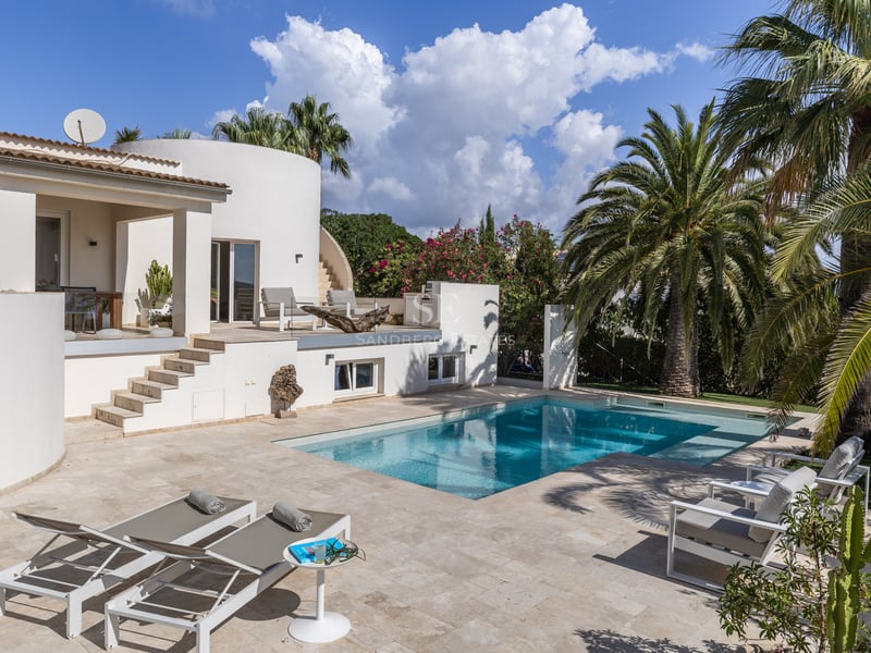 Villa blanche moderne avec piscine turquoise, terrasse en pierre naturelle et palmiers sous un ciel bleu éclatant.