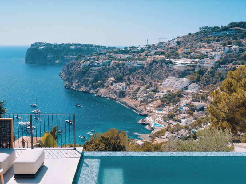 Terrasse de luxe avec piscine à débordement et fauteuils design surplombant une baie avec des yachts.