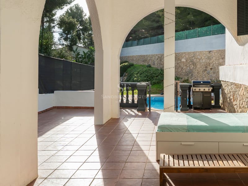 Terrasse couverte avec arches blanches, lit de repos, barbecue et vue sur une piscine ensoleillée.