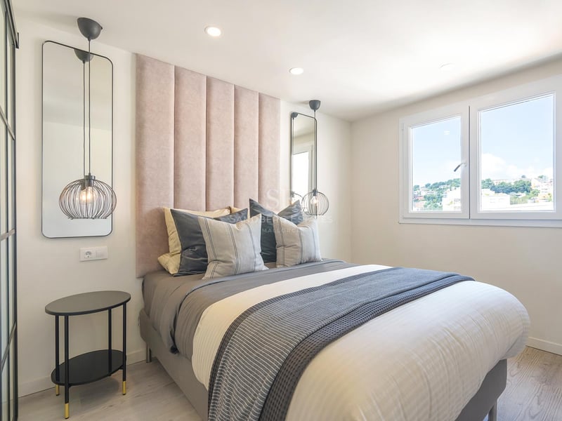 Chambre moderne avec tête de lit rose poudré, suspensions design, linge de lit gris et fenêtre lumineuse.
