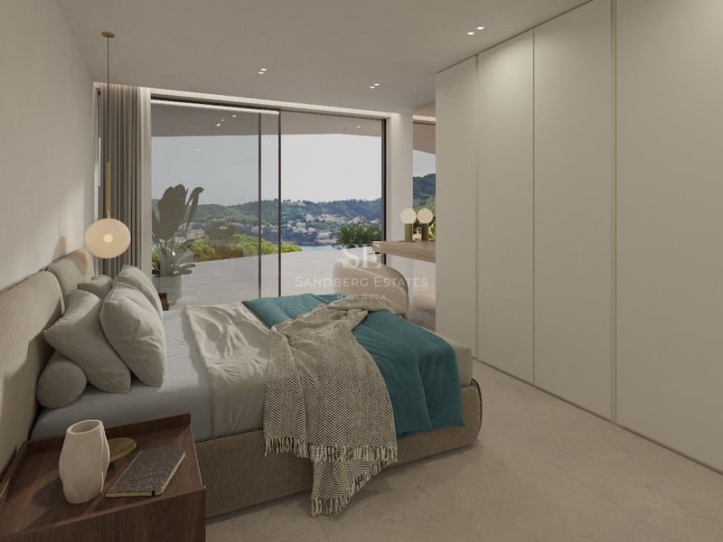 Chambre minimaliste avec lit rembourré, armoires encastrées et baies vitrées offrant une vue sur la mer et les montagnes.
