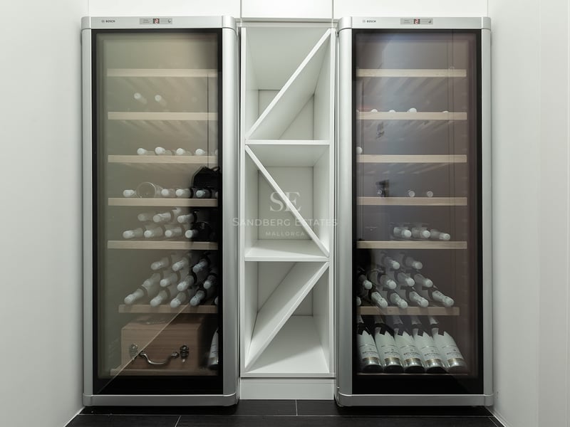 Deux élégantes caves à vin Bosch argentées encadrant une étagère à bouteilles blanche dans une pièce minimaliste.