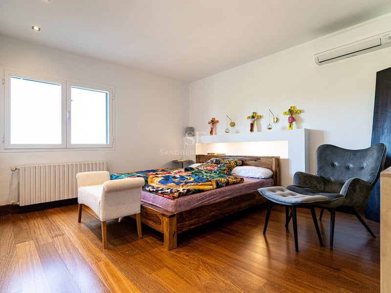 Chambre lumineuse avec parquet, cadre de lit en bois, fauteuil gris et fenêtre laissant entrer la lumière naturelle.