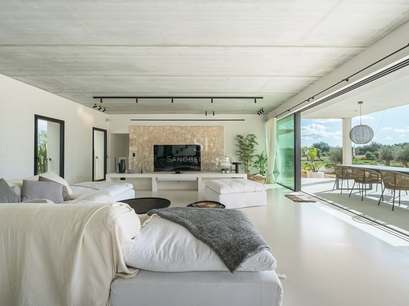 Salon moderne avec canapés blancs, plafond en béton, mur en pierre et baies vitrées ouvrant sur une terrasse.