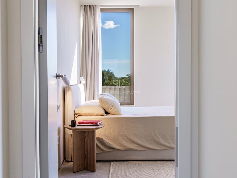 Vue à travers une porte sur une chambre lumineuse avec lit beige, table de chevet en bois et haute fenêtre.