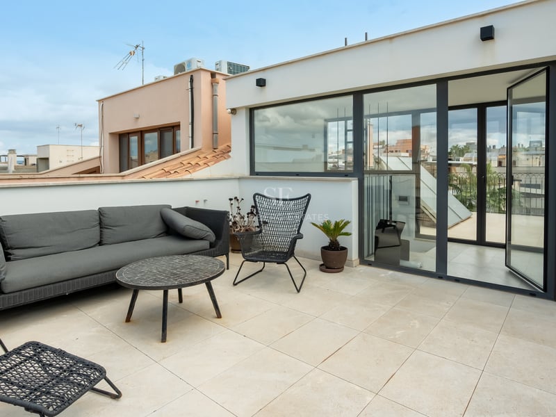 Terrasse sur le toit avec canapé gris, chaises design noires et grandes portes vitrées donnant sur la ville.