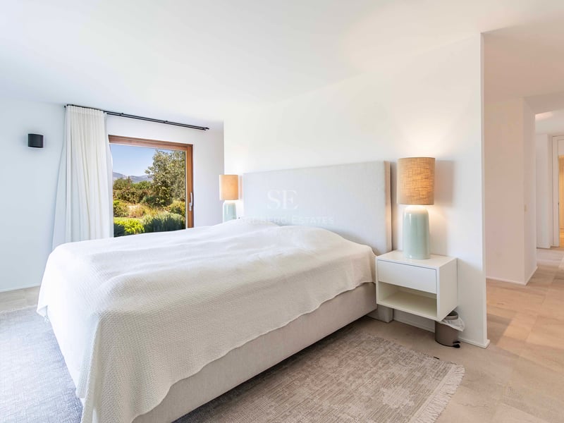 Chambre parentale lumineuse et moderne avec sol en pierre, lit rembourré et fenêtre donnant sur la verdure.