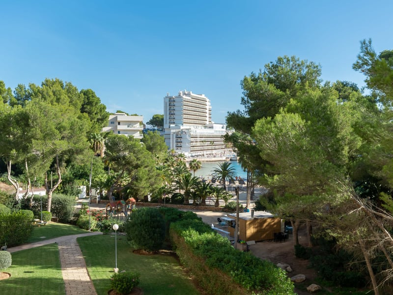 Jardin vert soigné avec des sentiers en pierre, des pins et un grand hôtel surplombant une baie sous un ciel bleu.