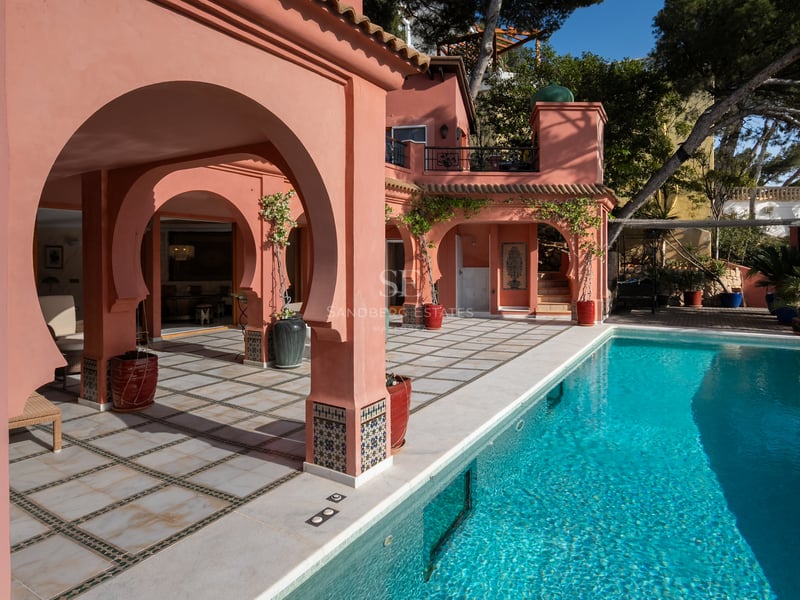 Piscine turquoise jouxtant une villa rose saumon avec arches mauresques et terrasse carrelée sous le soleil.