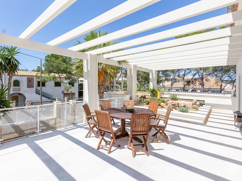 Terrasse ensoleillée avec une pergola blanche, une table ronde en bois et des chaises, entourée de palmiers.