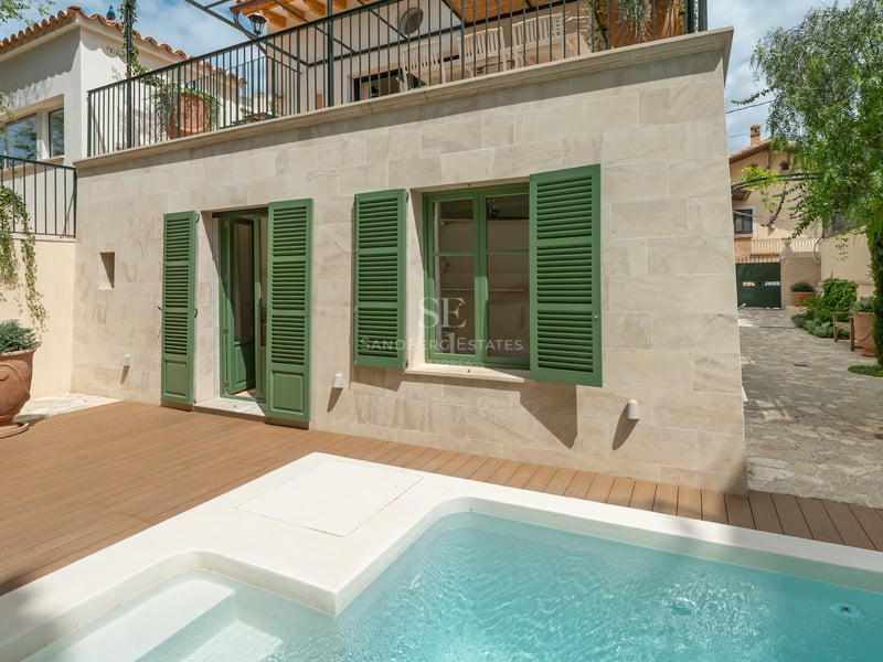Petite piscine moderne avec terrasse en bois à côté d'une façade en pierre claire et volets verts.
