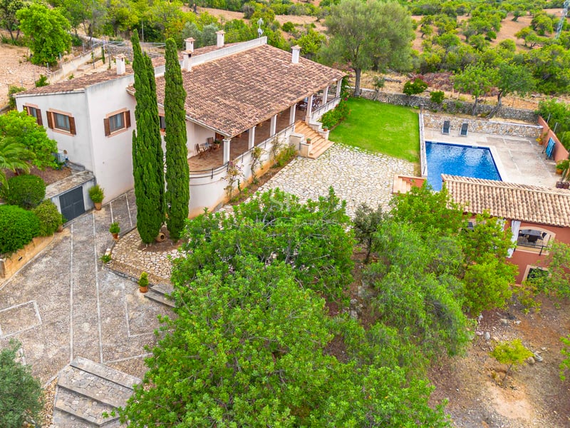 Vue en plongée d'une villa avec toit en terre cuite, cour en pierre, piscine bleue et aménagement paysager verdoyant.