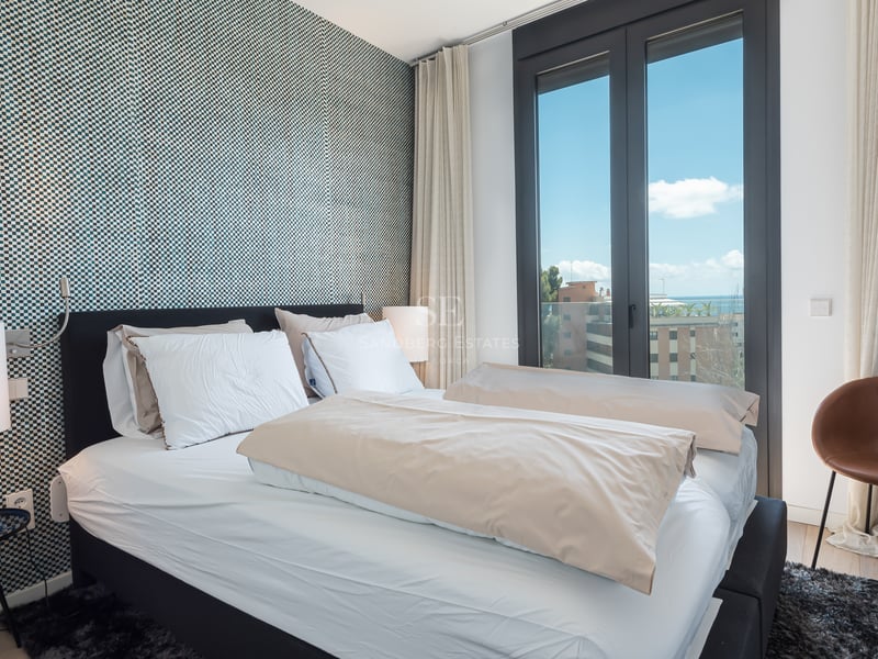 Chambre moderne avec mur tapissé bleu, lit blanc et grande fenêtre offrant une vue sur la mer et la ville.