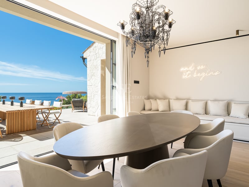 Salle à manger moderne avec table ovale sombre et chaises blanches ouvrant sur une terrasse ensoleillée surplombant la mer.