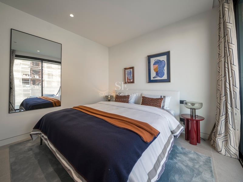 Chambre moderne lumineuse avec grand lit, literie bleu et orange, table rouge et un miroir reflétant une fenêtre.