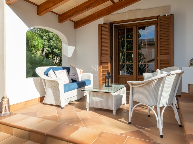 Mobilier en osier blanc sur une terrasse en terre cuite avec poutres en bois et vue sur le jardin.