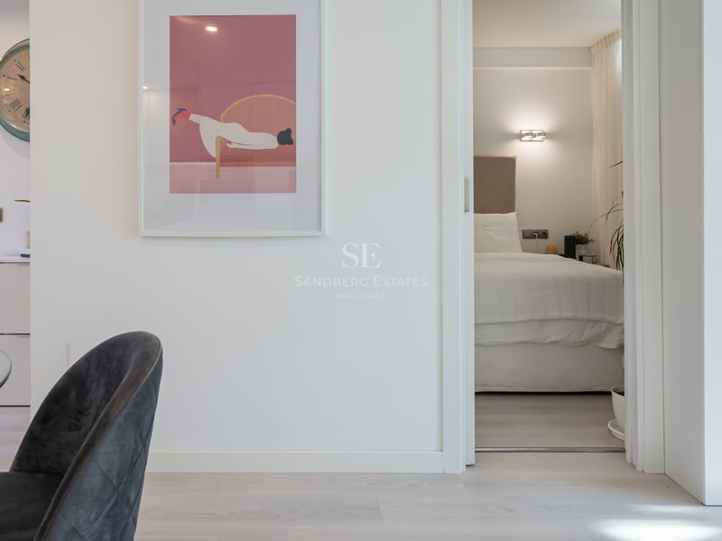 Vue à travers une porte ouverte sur une chambre minimaliste avec un grand lit, tête de lit beige et éclairage tamisé.