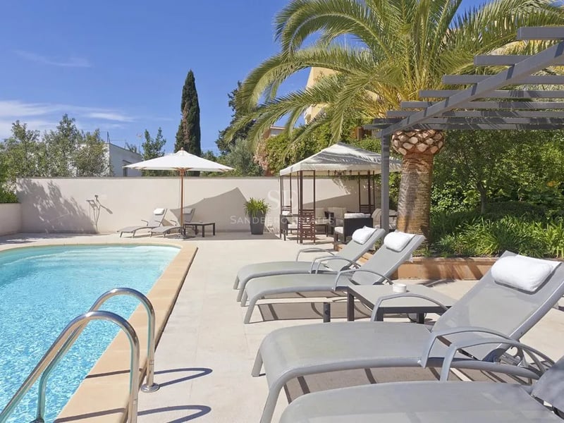 Piscine aux eaux turquoise entourée de transats gris, d'un palmier et d'une pergola sous un ciel bleu azur.