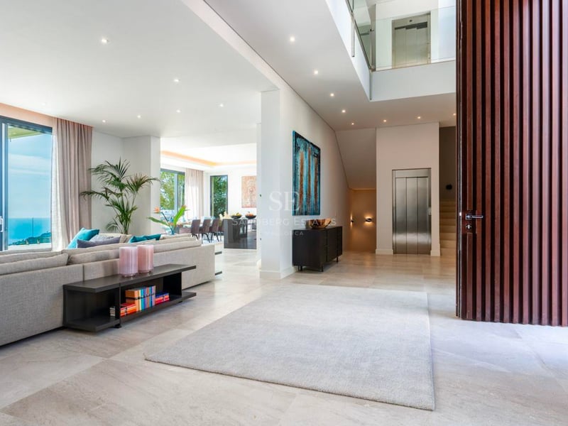 Salon moderne spacieux avec canapé gris, grandes baies vitrées avec vue sur mer et ascenseur intérieur.