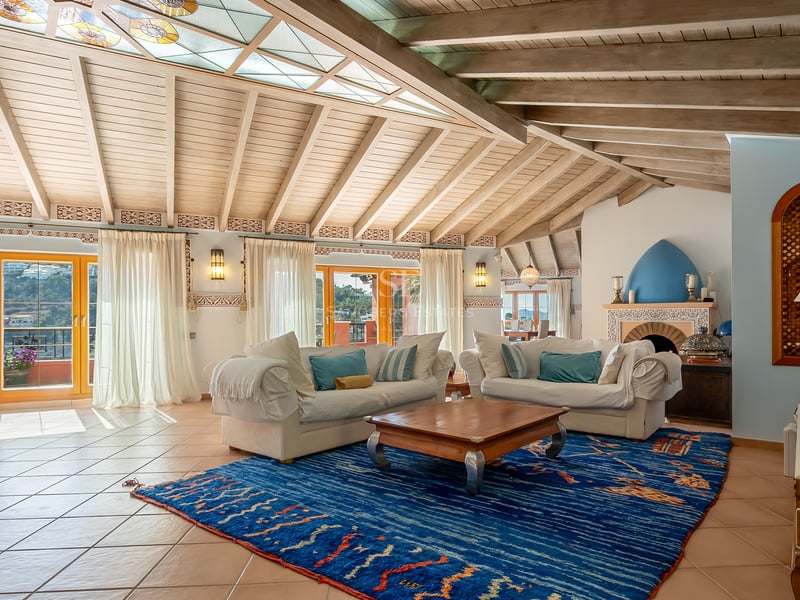 Grand salon avec poutres en bois apparentes, sol en terre cuite, canapés blancs et un grand tapis bleu à motifs.