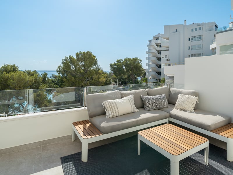Terrasse sur le toit avec canapé gris en L, table en bois et garde-corps en verre surplombant des pins et la mer.